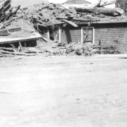 View of a shingle-sided house covered with a tree trunk, mud, and debris after a flood on the Arkansas River in Pueblo, Colorado. Multi-story brick buildings are in the distance.