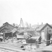 View of buildings and shacks moved and destroyed by the flood waters of the Arkansas River in Pueblo, Colorado. Debris, a boxcar, lumber, and utility poles are scattered among the buildings. Smoke stacks and steeples are in the distance.