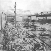 Cast iron cooking stoves are piled up against the side of the brick Holmes Hardware Store at 400 S. Union Avenue near railroad tracks after the Arkansas River flood,  in Pueblo, Colorado. Wreckage and debris are piled up near a viaduct.  Wooden framing braced up with boards is beside the track. Railroad boxcars are in the distance.