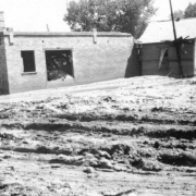 View of a small house that was carried by the flood waters of the Arkansas River and came to rest against the side of a brick garage in Pueblo, Colorado. The garage has damaged walls. Two men pose beside the garage. Ruts in the deep mud are near the buildings.