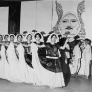 Teenage boys and girls, members of the Ballet Chicano de Aztlán, a Chicano dance troop and part of the Crusade for Justice, dance in Mexican costumes on a stage, in front of a painted drop in Denver, Colorado. The girls wear print dresses decorated with lace and lace mantillas. The boys wear white pants and shirts, one boy holds a candle.