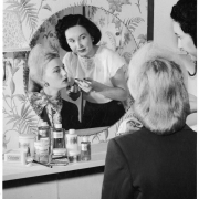 A woman applies lip gloss to a model posed in front of a mirror in probably Denver, Colorado. The model wears veil in her hair and scarf around her neck.  Bottles, jars, and boxes of cosmetics sit on a table, labels read: "Tributa's Studios." Shows floral wallpaper and a sconce.
