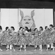 Teenage boys and girls, members of the Ballet Chicano de Aztlán, a Chicano dance troop and part of the Crusade for Justice, dance in Mexican costumes on a stage, in front of a painted drop in Denver, Colorado. The boys stand behind the girls, they wear headbands, the girls wear print dresses with full sleeves and  pleated, flared skirts.