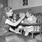 Interior view of toddler eating area, Pueblo Day Care Center (day  nursery), Pueblo, Colorado, shows a woman spoon-feeding three toddlers in high chairs. There are children wearing bibs, and glasses of milk and plates of food on trays.