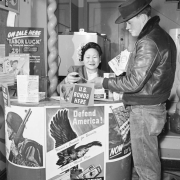 View of a war bonds promotion in a Denver, Colorado theater lobby.  A Japanese woman shows a man a brochure; signs read: "U.S. Bonds Here," "Defend America," and "On Sale Here - Tabor Luck by Caroline Bancroft."
