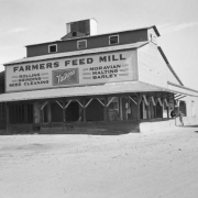 View of "Farmers Feed Mill," at 5870 Wadsworth Boulevard in Denver, Colorado; signs read "Feed Nutrena," and "Rolling Grinding Seed Cleaning Moravian Malting Barley." The building has a pitched monitor roof and a porch.