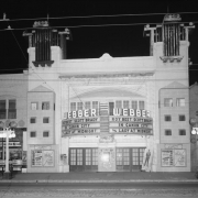 View of the Webber Theater, at 119 South Broadway Street in the Speer neighborhood of Denver, Colorado. The marquee reads: "Roy Best and Scott Brady in Canon City and Lady at Midnight." The building has a curved marquee and art deco style towers. Cast iron street lamps, bicycles, and storefronts are to the sides.