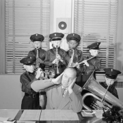 Boys in the Denver Junior Police Band pose with clarinets, a flute, a trumpet, and a bass cornet in Denver, Colorado. They wear uniforms and billed caps. A telephone is on a desk; the director covers his ears.