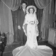 A bride and groom pose for a wedding studio portrait in (probably) Denver, Colorado. The bride's wedding dress has a puddle train; she wears a wide brimmed hat and carries a trailing orchid bouquet. The man wears a suit and tie.