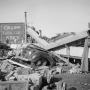 View of tornado damage to the Safeway grocery store in Julesburg (Sedgewick County), Colorado; shows a Ford sedan crushed under beams. People walk among debris; lettering reads: "Collins Furniture Store."