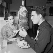 A man and woman eat sandwiches in (probably) Denver, Colorado; a baby hangs in a canvas baby carrier (cradleboard) from a hook on the wall.
