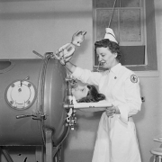 A Red Cross nurse talks to a woman in an iron lung (respirator) at Denver General Hospital (Denver Health Medical Center) in Denver, Colorado. Her uniform has a patch that reads: "American Red Cross," and she holds a toy stuffed rabbit.
