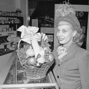 A woman poses with a basket of apples and grapes at the York Food Mart at 2021 East Colfax Avenue in Denver, Colorado. She wears a hat with feathers and a veil.