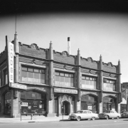 View of James Motor Company at 1278 Lincoln Street (at 13th / Thirteenth) in Denver, Colorado; features include stone gothic arches, stepped pediments with shield motifs and spindles. Signs read: "Dodge," "Plymouth," and "Parts."