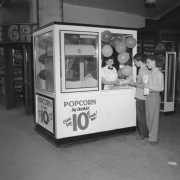 A teenaged boy and girl purchase popcorn at a stand festooned with balloons in (probably) Denver, Colorado. A sign reads: "Popcorn by Dunkin' 10 cents a Bag," a scale and shelves of liquor bottles are in a nearby concession; a neon sign reads: "Louie Gr...."