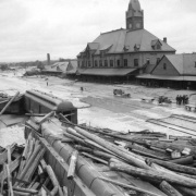 View of Union Depot in the aftermath of the 1921 flood in Pueblo, Colorado, shows debris and destruction. Piles of timber and beams are stacked against overturned railroad passenger cars. Mud and debris from the receded flood waters extends to the railroad station platform of Union Depot. Empty and loaded railroad baggage carts are next to a group of  men.