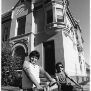 A boy and a girl (in sunglasses) pose on bicycles at the corner of 27th (Twenty Seventh) and Stout Streets in the Curtis Park Neighborhood of Denver, Colorado. A stone apartment building has corbeled brick, dentils, a corner turret, an arched window, and carved stone ornaments.
