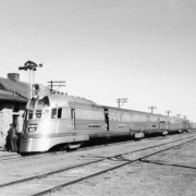 View of the Pioneer Zephyr  9900 running as Colorado and Southern Railway train 32 stopped at the railroad station in Longmont (Boulder County), Colorado. The streamlined art-deco style diesel locomotive and cars are constructed of stainless steel. The consist has a post office car, baggage car and passenger cars. Lettering on the train reads: "Burlington Route."