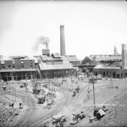 View of Globe Smelter at 52nd (Fifty-second), Logan, and Washington Streets in Denver, Colorado. Men work in the yard. Shows brick masonry buildings, furnaces, tall smelter chimneys, ladders, tailing piles, and carts. Several large ore crucibles sit in carts on the narrow gauge tracks of the Denver & Rio Grande Railroad.