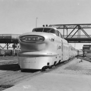 View of the General Motors experimental streamliner the Aerotrain led by a LWT12 locomotive at Union Station in Denver, Colorado.  The streamlined locomotive has four headlights. A steel viaduct and a flour mill are in the distance.