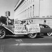 Walter Johnson, candidate for governor of Colorado sits in an open antique touring car with an unidentified man and boy on the corner of Fourteenth Avenue and Bannock Street in Denver, Colorado. Signs on the automobile read: "Walter Johnson for Governer [sic]." and "To Guarantee Labor's Rights Vote in the Primary Sept. 12. The Denver Public Library (Carnegie) is in the distance.