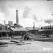 View of the work yard at the Globe Smelter at 52nd (Fifty-second), Logan, and Washington Streets in Denver, Colorado. A worker pushes an ore cart on the Denver & Rio Grande narrow gauge railroad tracks past a pile of loose rails. Shows brick masonry buildings. Men work under the eaves by the smelter. Chimneys belch smoke.