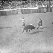 The bullfight on August 24 or 25, 1895, takes place in the bullring built at the race track in Gillett, Colorado, for the occasion. Joe Wolfe was the organizer of the event which caused a scandal because of its cruelty to animals. A bullfighter waves his cape at the bull, who paws the ground, while the matador looks on. The mounted picadors carry lances; one of the horsemen is seven-foot-tall, long-haired Charlie Meadows, Joe Wolfe's partner. Spectators are in the stands behind the wood fence.