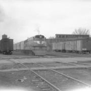 View of Chicago, Milwaukee, St. Paul, and Pacific Railroad Company streamliner "Hiawatha" led by steam locomotive #1 at a railroad crossing in Perry (Dane County), Wisconsin. Shows a coaling tower, an abandoned building and a railroad freight car on wooden blocks. A railroad signal is near the tracks.