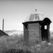 View of Finntown mining district's dilapidated structures in Stray Horse Gulch, east of Leadville (Lake County), Colorado. A small six-sided structure, possibly an outhouse, sits on a grassy slope in the foreground and is made of corrugated metal siding and has a faceted roof with finial ball, and a bracketed arched doorway. There are wood frame structures in the background and a tall smokestack on the left.