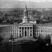 View of the Denver City and County Building at 1437 Bannock Street, in downtown Denver, Colorado; classic columns and entablature flank a pedimented entrance. A cupola / clock tower tops the curved facade; buildings, Front Range mountains and Mount Evans are in the distance.