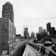 View of Broadway Street and viaduct in the Curtis Park Neighborhood of Denver, Colorado. Shows the former McPhee-McGinnity paint factory building and clock tower; downtown office buildings and skyscrapers are in the distance, and traffic is in the street. Letters read: "Butcher Block III Restaurant."