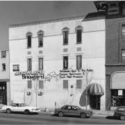 View of businesses at 12 Broadway Street in the Baker Neighborhood of Denver, Colorado. Brick and stucco buildings have awnings and signs: "Snowbird Designer Sportswear," and "Fair Exchange." Sedan automobiles are parked on the street.