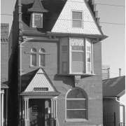 View of a three story brick house at 2418 Stout Street in the Five Points neighborhood of Denver, Colorado. The house has eclectic architectural elements that include arched windows with voussoirs, bargeboard gables, corbiestep sides, plaster relief details, and a dormer. A sign in the yard reads: "Offered by Marsh & Associates, 628-5474, For Rent."