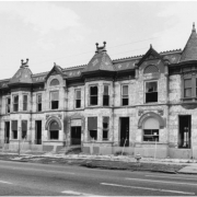 View of a two story stone row house at 2700 to 2714 Stout Street in the Five Points neighborhood of Denver, Colorado. The dilapidated house has High Victorian Gothic elements that include a corner turret, finials and a mansard roof. Shows missing doors and windows. A fence surrounds the property.