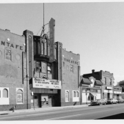View of the Aztlan Theatre at 974 Santa Fe Drive in the Lincoln Park neighborhood of Denver, Colorado. The brick building has Art Deco elements, a stepped front and low relief designs. Signs read "Aztlan" and "Santa Fe Theatre." The marquee reads "Hombres De Ti[e]rra Caliente," "Guerrillero Del Norte," "Gerado Reyes," "Karate," and "Live Boxing, Oct. 19 Fri." Shows shops and commercial facilities beside the theater. Automobiles are parked along the street.