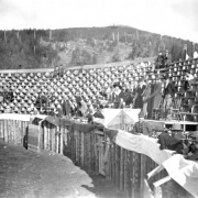 The bullfight on August 24 or 25, 1895, in the wood bullring built at the race track in Gillett, Colorado, is about to begin. Joe Wolfe, the organizer of the event and "Master of the Fiesta," wearing a Mexican sombrero, a velvet suit with silver buttons and gloves, stands in a decorated box in the stands, with four men behind him. People around look toward the bullring; there are many empty seats in the arena. The event caused a scandal because of its cruelty to animals.