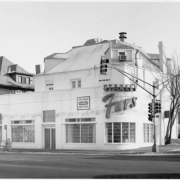 View of a business building at Colfax Avenue and Franklin Street in the Capitol Hill Neighborhood of Denver, Colorado. A storefront with a curved corner has been built onto a residence; a neon sign reads: "Stanley Furs." "Joe's Hair Affair" is next door.