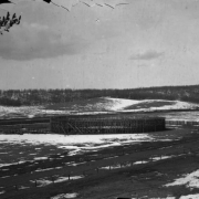The wood bullfight ring used for the Mexican bullfight on August 24 and 25, 1895, within the racetrack at Gillett, Colorado, is empty; snow is on the ground; trees on the hills behind are leafless. Joe Wolfe was the organizer of the event which caused a scandal because of its cruelty to animals.