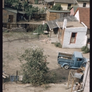 View of a pickup truck parked in a dirt backyard probably in the Sun Valley neighborhood, Denver, Colorado. Shows stuccoed brick houses, wooden fences and sheds and garages.