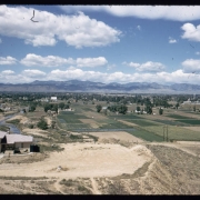 View of Arvada, Colorado, from Inspiration Point near 50th Avenue and Gray Street. Shows Clear Creek, the Wadsworth Drive-In Theater, the Arvada water tower, a two story stuccoed house, agricultural fields, and a bridge. Front Range mountains are in the distance.