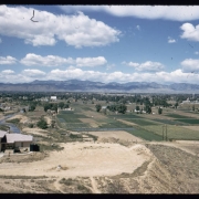 View of Arvada, Colorado, from Inspiration Point near 50th Avenue and Gray Street. Shows Clear Creek, the Wadsworth Drive-In Theater, the Arvada water tower, a two story stuccoed house, agricultural fields, and a bridge. Front Range mountains are in the distance.