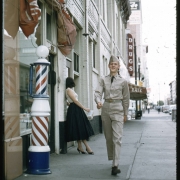 A young soldier in uniform walks down a street in the central Business District in Denver, Colorado. Shows a barber shop, a barber pole, and a sign for a drugstore. A woman leans against the wall of a hotel. She wears a blouse, skirt and high heels and carries a square plastic acrylic purse.