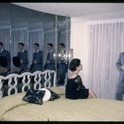 A man and a woman pose in a bedroom of a show unit at the Green Mountain Townhouses in Lakewood, Colorado. The room has a Queen sized bed with mirrors behind the headboard. Chandeliers that hang from chains hang over bedside tables. The woman wears a dark dress, has her hair pulled back in chignon and sits on the bed. The man wears a suit and tie and stands in front of a glass door covered with curtains.
