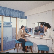 View of the kitchen area and breakfast bar in a unit at the Green Mountain Townhouses in Lakewood, Colorado. A glass patio door looks out on a brick patio with a table, an umbrella and chairs. A man sits at the breakfast bar on a stool. Women stand near the breakfast bar and a stove. Shows a refrigerator and cupboards. A valance is over the door.