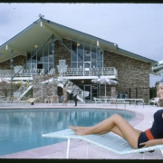 View of the clubhouse and swimming pool at the Green Mountain Townhouses in Lakewood, Colorado. The modern brick glass and steel building has an irregularly shaped pitch roof, wall to ceiling glass windows, balconies and external staircases. A woman sits in a chaise lounge near the pool. Shows outdoor tables, chairs and aluminum umbrellas.