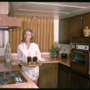 A woman stands in the kitchen of a model town home at the Green Mountain Townhouses in Lakewood, Colorado. She holds a tray of glasses and wears a white shirt, pink slacks, and an apron. Shows a built-in, wood grained, gas stove inset into a pink counter, an exhaust hood, a bronzed brown, built-in oven inset into a wooden enclosure and wood kitchen cabinets. Vases, artificial flowers and nic-nacs decorate the counters.