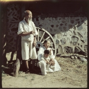 An elderly Korean man, his wife and a young boy pose near a stone and cement building in South Korea. The man wears linen knee length pants (paji) and a loose shirt with 3/4 length sleeves. He is balding and has a beard and long mustache. He smokes a long, bamboo and metal traditional Korean pipe. The woman sits beside the man and wears a white linen dress (hanbock). The child wears a sweater.