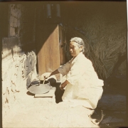An elderly Korean woman kneels beside an outdoor hearth and stirs an iron pot in South Korea. The lid of the pot is beside the pot. The woman wears a loose fitting white linen traditional dress (hanbock). She wears her gray hair pulled back.