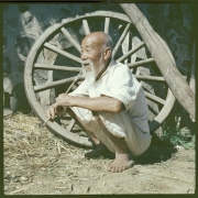 An elderly Korean man squats near a large wagon wheel that leans against a stone and cement wall in South Korea. He wears linen knee length pants (paji) and a loose shirt with 3/4 length sleeves. He is balding and has a beard and long mustache. He smokes a long, bamboo and metal traditional Korean pipe.