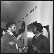 Two African American men, who include painter and sculptor Bob Ragland, stand and chat at an art show at the Denver Art Museum in Denver, Colorado. Mr. Ragland wears a leather jacket and dark glasses, his hair is in a short afro and he has a beard and mustache. The other man wears a sports coat and a scarf. A sign on the wall reads: "Contemporary Black Artists Denver, '73."